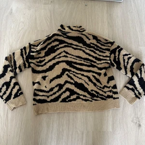 Beige och svart zebramönstrad polotröja - Snygg stickad polotröja med beige och svart zebramönster. Tröjan har lång ärm, ribbade muddar och hög krage.