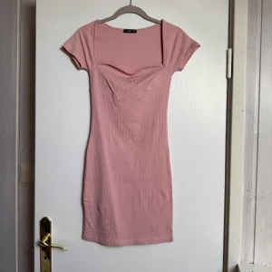 Rosa ribbad kortklänning Dilvin - Supersöt rosa kortklänning från Dilvin med ribbad struktur och smickrande drapering vid halsen. Klänningen har korta ärmar och en figurnära passform som framhäver siluetten. Perfekt för dig som gillar enkel men snygg stil. Oanvänd.