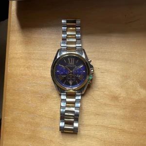 Michael Kors klocka i silver och guld - Stilren Michael Kors klocka med länkarmband i silver och guld. Urtavlan är svart med blå detaljer och romerska siffror, samt flera små urtavlor. Klockan har guldiga knappar på sidan och är tillverkad i rostfritt stål.