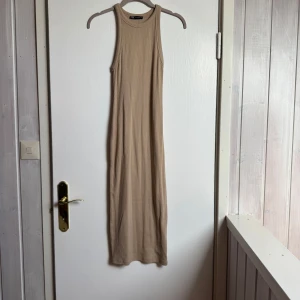 Beige ribbad långklänning från Zara - Säljer en beige ribbad långklänning från Zara med ärmlös design och rund hals. Klänningen har en slim passform och är tillverkad i mjuk bomull som sitter skönt mot kroppen. Perfekt för dig som gillar stilrena och enkla plagg. Oanvänd.