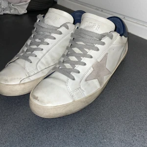 Vita sneakers Golden Goose med stjärna - Säljer ett par vita sneakers från Golden Goose med grå mockadetalj i form av en stjärna på sidan och blå hälkappa. Skorna har grå snörning och platt sula. Klassisk och clean look med coola detaljer som GGDB/SSTAR-tryck på sidan.