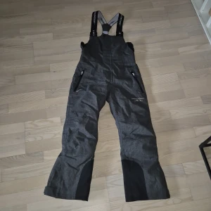 Grå täckbyxor/skidbyxor från Swedemount str 140 - Säljer ett par grå täckbyxor från Swedemount med svarta förstärkningar vid bensluten och justerbara hängslen. Byxorna är vattentäta, vindtäta och andas, perfekta för vinterns alla äventyr. Utrustade med dragkedjor på fickorna och hög midja för extra skydd. Nypris 999:-