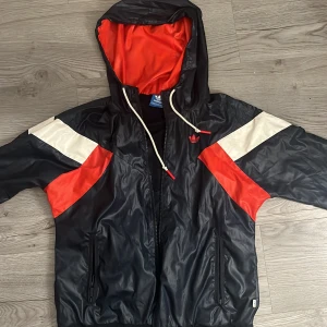 Svart och röd vindjacka från Adidas - Snygg vindjacka från Adidas i svart med röda och vita detaljer. Jackan har huva med dragsko, dragkedja framtill och två fickor. Insidan är fodrad med mesh och jackan har Adidas logga på bröstet. Perfekt för dig som gillar sportig stil.