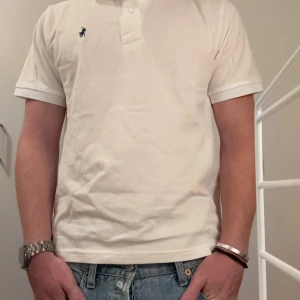 Vit pikétröja från Polo Ralph Lauren - Klassisk vit pikétröja från Polo Ralph Lauren i bomull. Tröjan har korta ärmar, krage och knäppning framtill samt den ikoniska broderade loggan på bröstet. Classic fit-modell som passar till jeans eller shorts.