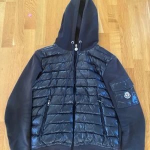 Svart Moncler pufferjacka med huva - Snygg svart pufferjacka från Moncler i storlek S. Jackan har glansig quiltad front, mjuka ärmar och huva med dragsko. Ficka med Moncler-logga på ärmen och dragkedjor framtill. Perfekt för dig som vill ha en stilren och sportig look.