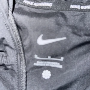 Svart Nike vindjacka med huva - Säljer en svart Nike vindjacka i storlek M. Jackan har huva med dragsko, hel dragkedja framtill och Nike-logga på bröstet. Lätt och smidig modell som passar perfekt för löpning eller andra utomhusaktiviteter. Materialet är tunt och känns sportigt.