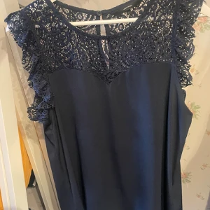 Marinblå spets blus från Vero Moda - Supersnygg marinblå blus från Vero Moda med vacker spets upptill och ärmlös design. Klänningen har rund halsringning, knäppning bak i nacken och ett stilrent, rakt fall. Perfekt för dig som vill ha en elegant look med extra detalj vid axlarna.