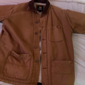 Brun workwearjacka från Dickies L - Säljer en brun workwearjacka från Dickies i storlek L. Jackan har stora fickor framtill, bröstficka med logga och brunt kragparti. Insidan är fodrad med mjuk teddy i offwhite. Stängs med knappar och har en klassisk, rak passform.