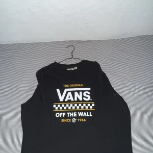 Svart långärmad Vans tröja XL junior  - Svart långärmad tröja från Vans i storlek XL junior med stort tryck framtill i vitt och gult. Klassisk rund halsringning och schackrutigt mönster på bröstet. Perfekt för dig som gillar streetwear och vill sticka ut med en ikonisk look.