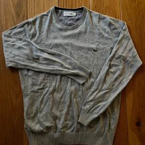Grå stickad tröja från Lacoste - Snygg grå stickad tröja från Lacoste med klassisk rund halsringning och den ikoniska krokodilloggan på bröstet. Tröjan har ribbade muddar vid ärmslut och nederkant, perfekt för lager-på-lager och chill stil.