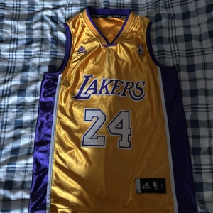 Gul Lakers baskettröja Adidas M - Snygg gul och lila Los Angeles Lakers baskettröja från Adidas med nummer 24 på bröstet. Tröjan är ärmlös och har klassisk V-ringning, tillverkad i glansigt syntetmaterial. Lakers-logga och NBA-märke framtill. Perfekt för fans!