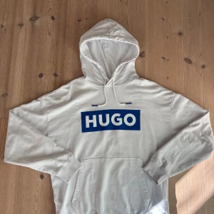 Hugo hoodie vit/blå - Vit hoodie från HUGO med stor blå logga framtill. Klassisk känguruficka, dragsko i huvan med blå detaljer och mjukt bomullsmaterial. Perfekt för dig som gillar stilren streetwear med tydlig branding. Ny utan prislapp och är helt ren samt inga skador.