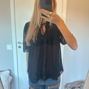 Svart blus med prickig struktur KappAhl - Säljer en svart blus från KappAhl med transparent tyg och prickig struktur. Blusen har korta ärmar, smockdetaljer upptill och knytning vid halsen. Luftig och flowy passform, perfekt för en trendig look.