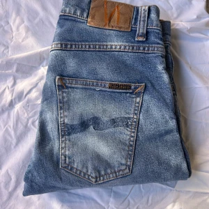 Nudie Jeans Grim Tim blå jeans 29/32 - Säljer ett par Nudie Jeans Grim Tim. Klassiska blå jeans med snyggt slitna detaljer, orange sömmar och knappgylf. Tillverkade i 99% bomull och 1% elastan för lite stretch. Perfekta för dig som gillar en tidlös och avslappnad look.