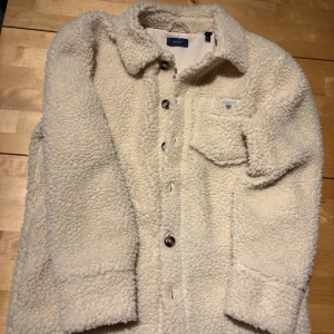 Vit teddyjacka från Gant, 146/152 - Mysig och trendig vit teddyjacka från Gant med klassisk krage, bröstficka med broderad logga och knäppning framtill. Jackan är fluffig och har lång ärm, perfekt för kyliga dagar. Passar dig som gillar en avslappnad och cool stil.