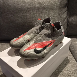 Nike Phantom Vision Elite fotbollsskor - Säljer ett par grå Nike Phantom Vision Elite fotbollsskor med röda och svarta detaljer. Skorna har en strumpliknande passform, diskret snörning och mönstrad ovandel i syntetmaterial för extra grepp. Sulan har fasta dobbar för spel på gräs. Riktigt snygga och moderna skor för dig som vill sticka ut på planen.
