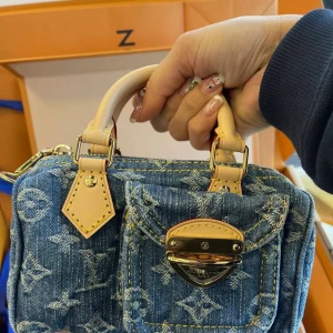 Louis Vuitton denim handväska - Superfin liten handväska från Louis Vuitton i blått denimtyg med ikoniskt monogrammönster. Väskan har ljusa skinnhandtag, guldfärgade metalldetaljer och dragkedja upptill. Framtill finns en ficka med snyggt spänne. Perfekt accessoar för att lyfta din outfit.