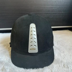 Svart ridhjälm från HaloRider - Snygg svart ridhjälm från HaloRider med rundad form och ventiler på sidorna. Framtill sitter en silverfärgad detalj med ventilationshål och HaloRider-logga. Ytan är slät och matt, och hjälmen har en klassisk look som passar till ridning.