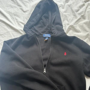 Svart hoodie från Polo Ralph Lauren - Svart hoodie från Polo Ralph Lauren med dragkedja och huva. Klassisk röd logga broderad på bröstet. Mjuk och skön bomull, perfekt för chill dagar. Långärmad modell med fickor framtill.