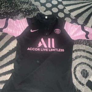 PSG x Nike svart och rosa fotbollströja - Snygg PSG fotbollströja från Nike i svart med rosa detaljer och tryck. Tröjan har korta ärmar i ljusrosa med svarta inslag och klubbmärke på bröstet. Materialet är lätt och andas, perfekt för träning eller match.