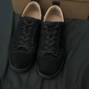 Svarta sneakers med röd sula - Säljer ett par svarta sneakers från Christian Louboutin med klassisk röd sula och svarta snören. Skorna har en stilren design med rund tå och är tillverkade i mocka och skinn. Insidan är ljusbeige vilket ger en snygg kontrast mot det svarta yttermaterialet. Skorna är använda ett par gånger och lite slitna men funkar fortfande 