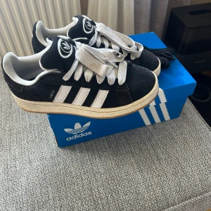Adidas Campus svarta sneakers mocka - Säljer ett par klassiska Adidas Campus sneakers i svart mocka med vita detaljer och de ikoniska tre ränderna på sidan. Skorna har vit sula, breda vita skosnören och Adidas logga på plös och häl. Perfekta för en stilren och sportig look.