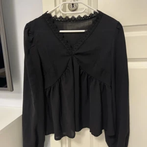 Svart blus med spets och v-ringning - Svart blus med v-ringad hals och dekorativ spetskant. Modellen har långa ärmar med lätt puff och rynk vid axlar och ärmslut. Blusen har en lös passform och volangdetaljer framtill som ger en romantisk vibe. Perfekt för dig som gillar stilrena och feminina plagg.