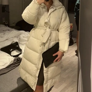 Vit lång pufferjacka med bälte - Säljer en lång, vit pufferjacka med bälte i midjan och stora, mysiga kanaler. Jackan har huva, knappar och dragkedja framtill samt långa ärmar. Perfekt för kalla dagar när du vill vara både varm och snygg. Säljs för halva priset ✨