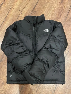 Svart pufferjacka från The North Face - Säljer en svart pufferjacka från The North Face med hög krage och dragkedja framtill. Jackan har en oversized look och är perfekt för kalla dagar. Klassisk logga på bröstet och ryggen. Fylld med dun och har två fickor med dragkedja.                     Knappt använd!!!!