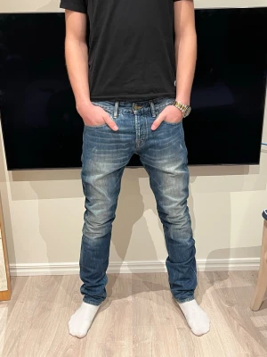 Scotch & Soda Ralston jeans blå - Säljer ett par blå Scotch & Soda Ralston jeans med snyggt slitna detaljer och klassisk femficksdesign. Jeansen har normal passform, raka ben och är tillverkade i bomull. Märkespatch på insidan och coola kontrastsömmar. Perfekt för dig som gillar en avslappnad stil.