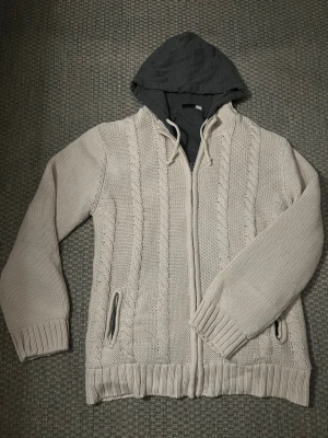 Beige stickad hoodie med dragkedja - Säljer en beige stickad hoodie med mörkgrå huva och snörning. Tröjan har dragkedja framtill, kabelstickat mönster och två fickor. Perfekt för kyliga dagar och riktigt mysig att ha på sig.