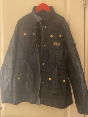 Svart  jacka från Barbour - Säljer en svart jacka från Barbour International med bälte i midjan, guldfärgade knappar och flera fickor. Jackan har hög krage, dragkedja och tryckknappar framtill samt Barbour-logga på bröstet. Perfekt för dig som vill ha en klassisk och snygg look.