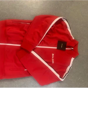 Röd Palm Angels zip hoodie - Palm Angels röd hoodie med vit dragkedja och vita ränder längs ärmarna. Klassisk streetwear-look med logga på bröstet och ribbade muddar. Perfekt för dig som vill sticka ut med en sportig vibe.