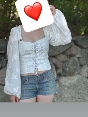 Vit korsettopp med snörning - Supersöt vit korsettopp med snörning framtill och puffiga långärmar. Toppen har ett diskret blommönster i tyget och är croppad med fyrkantig urringning. 