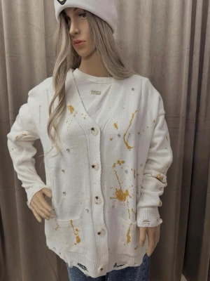 Vit kofta med guldstänk och pärldetaljer - Säljer en oversized vit stickad kofta med coola guldstänk och pärldetaljer. Koftan har knappar framtill, två fickor och slitna detaljer vid ärmslut och nederkant för en edgy look. Perfekt för dig som gillar unika plagg med streetkänsla.