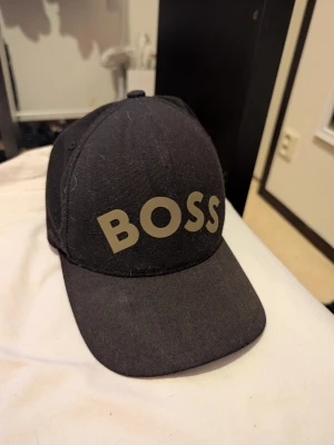Svart keps från BOSS med guldiga detaljer - Säljer en svart keps från BOSS med stor logga framtill och justerbart spänne bak i guld. Skärmen har ett unikt geometriskt mönster i guld som ger en snygg kontrast. Kepsen är i ett mjukt material och passar perfekt till streetwear-stilen.