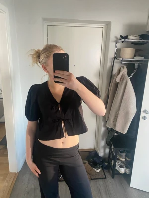 Svart blus med puffärm från Gina Tricot - Supersnygg svart blus från Gina Tricot med puffiga korta ärmar och två knytningar framtill. Croppad modell med v-ringning, perfekt till både jeans och kjol. Gjord i ett lätt bomullstyg som känns luftigt och skönt.