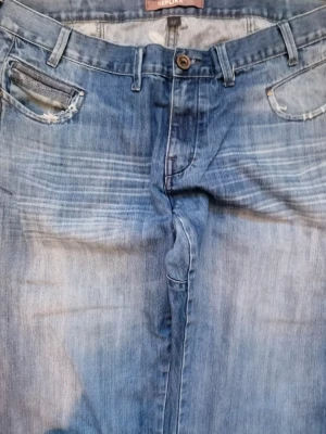 Blå jeans från Replika med broderi - Säljer ett par blå jeans från Replika med ljus tvätt och slitningar vid fickorna. Baksidan har stora broderade bokstäver och detaljerade bakfickor. Klassisk femficksmodell med raka ben och bälteshällor. Perfekta för dig som gillar streetwear och unika detaljer.