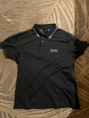 Svart piké från Hugo Boss - Snygg svart pikétröja från Hugo Boss med vit logga på bröstet och ryggen. Klassisk krage med vita ränder och knappar framtill. Tillverkad i mjuk bomull som känns skön mot huden. Perfekt till jeans eller shorts för en clean look.