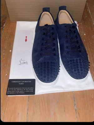 Mörkblå sneakers från Christian Louboutin - Säljer ett par mörkblå sneakers från Christian Louboutin i mocka med ikoniska nitar på tån och vit sula med röd undersida. Skorna har snörning och exklusiv design, perfekt för dig som vill sticka ut. Kommer med originalkartong, dustbag och äkthetskort. Hör av er vid frågor✅