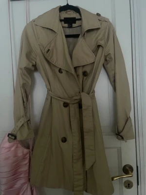 Beige trenchcoat från H&M - Snygg beige trenchcoat från H&M i klassisk modell med dubbelknäppning, bred krage och bälte i midjan. Jackan har långa ärmar med spännen vid ärmsluten och stora knappar framtill. Perfekt för vår och höst när du vill ha en stilren look.