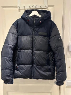 Mörkblå vinterjacka från Tommy Hilfiger - Snygg mörkblå pufferjacka från Tommy Hilfiger med huva och dragkedja framtill. Jackan har vattenavvisande material och värmande foder, samt logga på bröstet och ärmen. Perfekt för kalla dagar och har praktiska fickor med dragkedja.