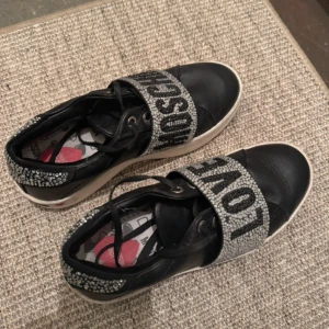 Love Moschino sneakers med strass och text - Snygga svarta sneakers från Love Moschino med breda band över foten med texten 'LOVE' och 'MOSCHINO' i silver. Dekorerade med glittrande strassdetaljer på hälen och bandet. Tjock vit sula med logga och hjärta på sidan.
