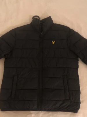 Svart pufferjacka från Lyle & Scott - Svart pufferjacka från Lyle & Scott med gul broderad logga på bröstet. Jackan har ståkrage, dragkedja framtill och två fickor. Den quiltade designen ger en schysst look och håller dig varm under kyliga dagar.