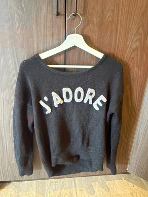 Svart stickad tröja med J'ADORE-tryck - Cool svart stickad tröja med stort vitt J'ADORE-tryck på bröstet. Tröjan har en rund halsringning och långa ärmar, perfekt för att ge din outfit en fransk vibe. Passar dig som gillar statement-plagg och vill ha något mjukt och bekvämt.
