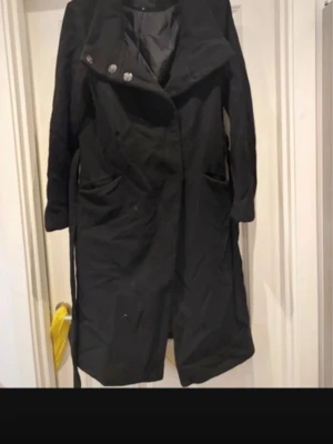 Svart lång trenchcoat med bälte - Säljer en svart trenchcoat med bälte i midjan och stora slag. Jackan har knappar framtill och två fickor. Lång modell som ger en clean och stilren look. Perfekt för dig som vill ha en klassisk och enkel jacka till höst eller vår.