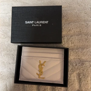 Vit Saint Laurent plånbok i skinn - Säljer en stilren vit plånbok från Saint Laurent Paris i äkta skinn. Plånboken har quiltad design, flera kortfack och ikonisk YSL-logga i guld framtill. Perfekt för dig som vill ha något lyxigt och snyggt i väskan.