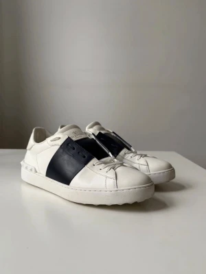Valentino Garavani vita sneakers - Säljer ett par vita Valentino Garavani sneakers med svart band över mitten och klassisk snörning. Skorna har rund tå, platt sula och är tillverkade i skinn. Medföljer extra skosnören med Valentino-logga. Perfekta för dig som gillar exklusiva och stilrena sneakers.