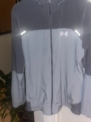 Grå vindjacka från Under Armour - Snygg tvåfärgad grå vindjacka från Under Armour med huva och hel dragkedja. Jackan har reflekterande detaljer, logga på bröstet och stort tryck på ryggen. Perfekt för utomhusaktiviteter och träning. Lätt material och praktiska fickor.