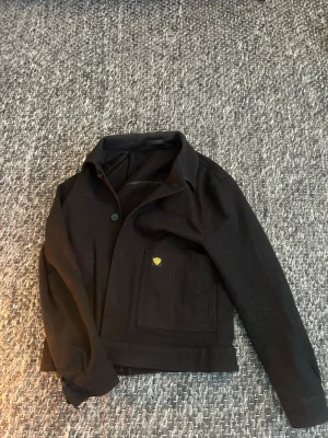 Svart overshirt med fickor - Säljer en svart overshirt med klassisk krage och stora framfickor. Jackan har knappstängning framtill och en liten gul detalj på ena fickan. Tillverkad i ett slitstarkt bomullstyg och har långa ärmar. Perfekt för lager på lager och enkel att styla.